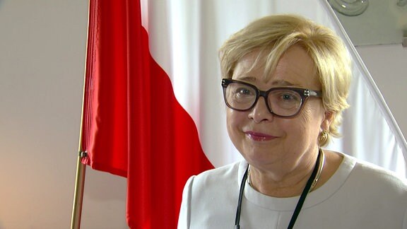 Blonde Frau mit Brille vor rot-weißer Fahne.