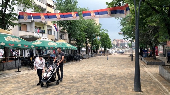 Ein Paar mit Kinderwagen in einer fast leeren Fußgängerzone. Über ihnen hängen mehrere serbische Flaggen.