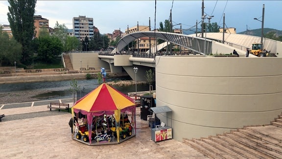 Blick auf eine moderne Brücke über einen Fluss. Im Vordergrund eine Uferpromenade mit einem bunten Kinderkarussel.
