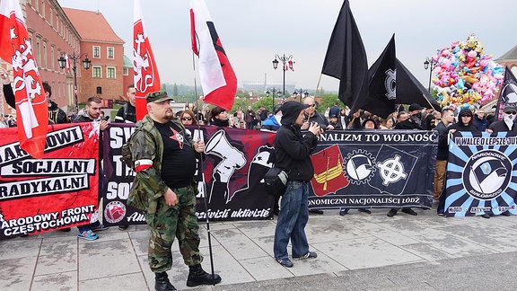Mann mit Tarnfleckuniform und polnischer Flagge, Dahinter vermummte Männer mit rechtsradikalen Transparenten.