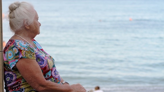 Alte Frau in buntem Hauskleid sitzt auf einer Mauer und schaut auf das Meer