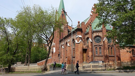 Rote Backsteinkirche. Davor Bäume und einzelne Besucher