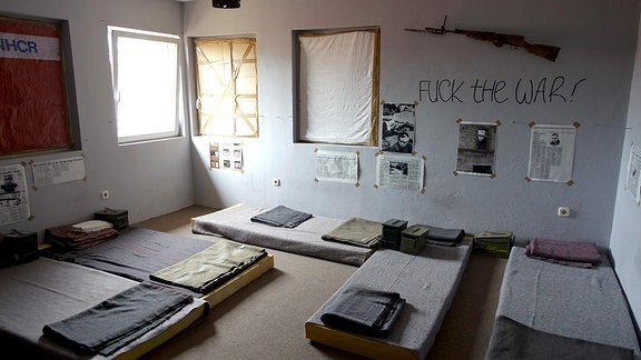 Karger Raum mit Matratzen auf dem Boden und Gewehr an der Wand. Daneben steht: „Fuck War“.