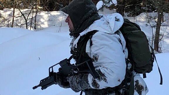 Ein Mann im Tarnanzug, Rucksack und einer Waffe in der Hand läuft durch den verschneiten Wald