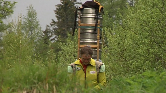 Mann in gelber Jacke trägt einen Stapel Bierfässer auf dem Rücken durch einen lichten Wald