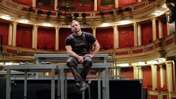 Mann in Arbeitskleidung sitzt auf einer Plattform in einem Konzertsaal.