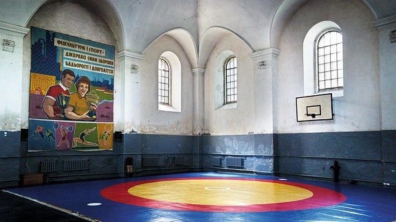 Eine Sporthalle mit bröckelndem Putz von innen. An der Wand ist ein Sportplakat mit kyrillischer Schrift. Auf dem Boden liegt eine Sportmatte.