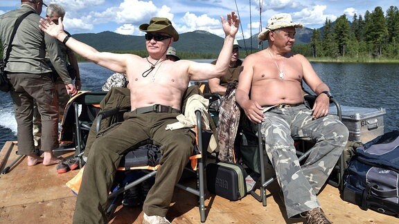 Der Russische Präsident Wladimir Putin und sein Verteidigungsminister Sergej Schoigu sitzen mit freiem Oberkörper am Bootssteeg.