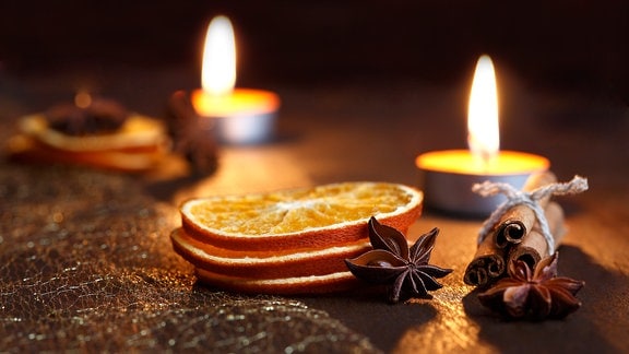 Zwei angezündete Teelichter, Zimtstangen, Anis und Orangenscheiben liegen auf einem dunklen Holz