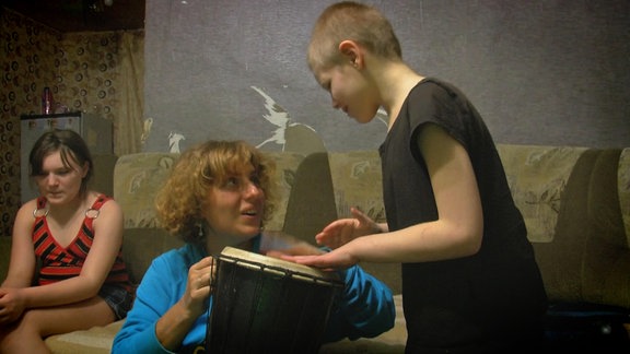 Eine Psychologin sitzt in einer heruntergekommen Wohnung in der Ukraine, nahe der Font, auf einem Sofa und spricht über die psyschichen Probleme der Kinder. Links von ihr sitzt ein kleines Mädchen und rechts von ihr ein junge mit Autismus.