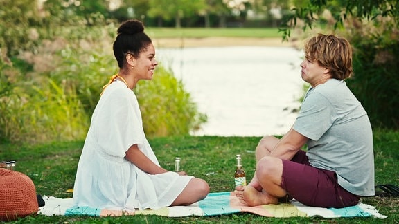 Eine junge Frau und ein junger Mann sitzen am Seeufer und picknicken