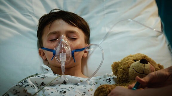 Ein kleiner junge liegt mit seinem Teddy im Krankenbett und muss künstlich beathmet werden