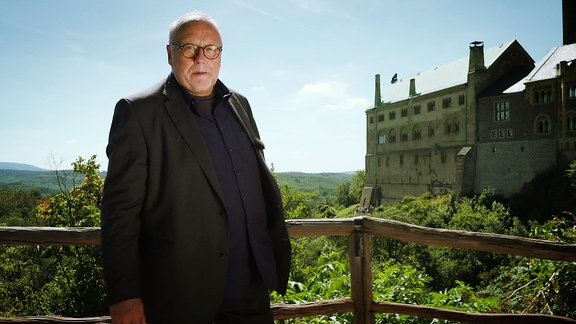 Ein älterer Herr steht an einem Aussichtspunkt. Im Hintergrund sinde Berge und eine Burg zusehen.