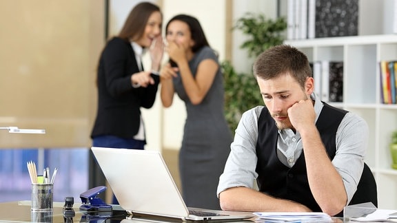 Two coworkers bullying a worried colleague that is sitting in his workplace at office