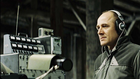 Stasi-Hauptmann Gerd Wiesler (Ulrich Mühe) sitzt in dem Kinofilm (Das Leben der Anderen) auf dem Dachboden eines Hauses und belauscht die Vorgänge in der Wohnung die er beschattet.