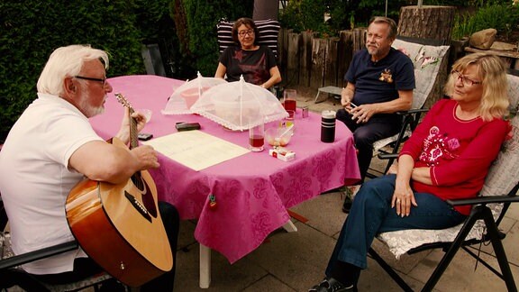 Vier Campingfreunde sitzten draussen an einem Tisch bei Kaffee und Kuchen. Einer der Freunde spielt Gitarre.