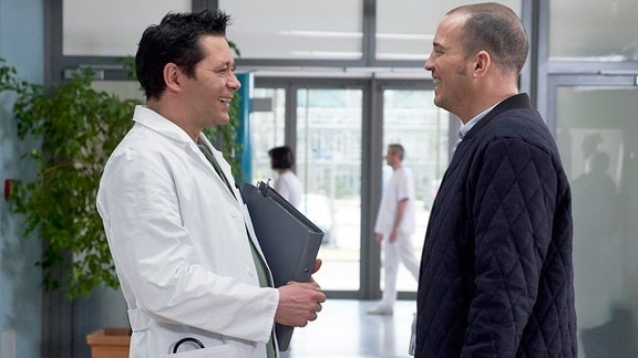 Ein Arzt und ein Mann stehen sich im Krankenhausflur lachend gegenüber