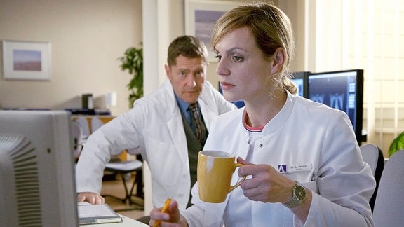 Dr. Lea Peters (Anja Nejarri) sitzt an einem Computer. Dr. Kaminski (Udo Schenk) schaut ihr über die Schultern.