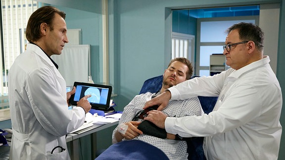 Dr. Martin Stein (Bernhard Bettermann, li.) und Hans-Peter Brenner (Michael Trischan, re.) sprechen mit einem Patienten über dessen Schulterverletzung.