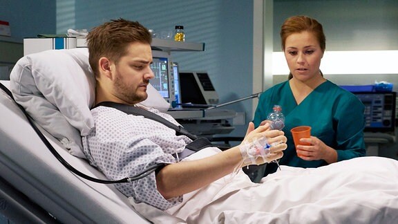 Schwester Julia (Sarah Tkotsch) sitzt am Bett eines Patienten. Dieser versucht seine Hand zu bewegen.