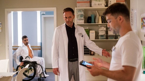 Kai Hoffmann steht wütend vor Pfleger Kris, der gerade auf ein Tablet schaut und Informationen kontrolliert, im Schwesternzimmer. Dr. Demir fährt mit seinem Rollstuhl auf die beiden zu.