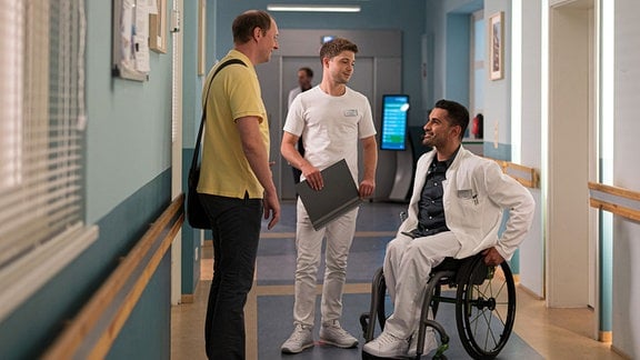 Im Gang der Sachsenklinik stehen Herr Meyer, Pfleger Kris und Dr. Demir sitzt im Rollstuhl.