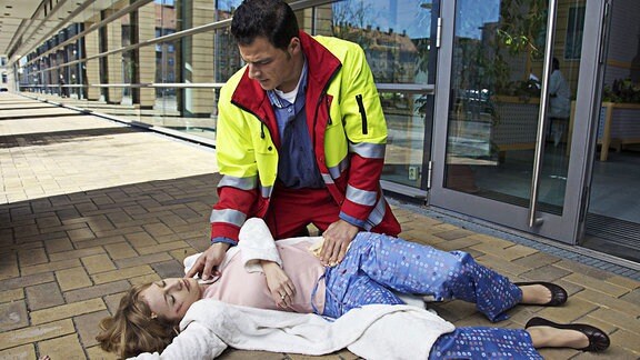 Cornelia Weiß liegt blutend auf dem Boden, ihre Operationsnarbe ist aufgegangen. Der Notarzt Raphael König behandelt sie.