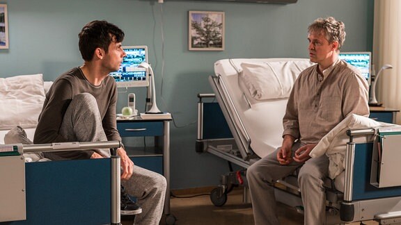Matthias und Marco sitzen in grauen Schlafanzügen einander gegenüber, jeder auf einem Krankenbett.