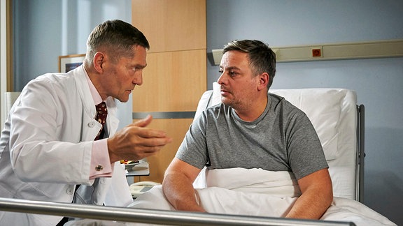 Dr. Kaminski (Udo Schenk, li.) erklärt Joachim Bindel (Rüdiger Klink, re.) am Krankenbett den Ablauf einer OP.