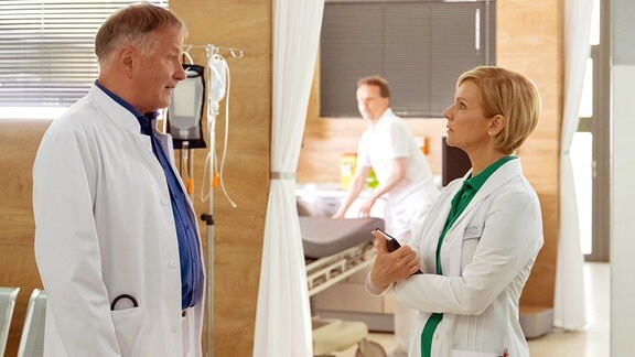 Zwei Mediziner auf Station diskutieren einen Fall. Unscharf im Hintergrund ein Pfleger im Krankenzimmer.