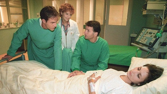 Ein Bekannter der Patientin und zwei Ärzte am Krankenbett.