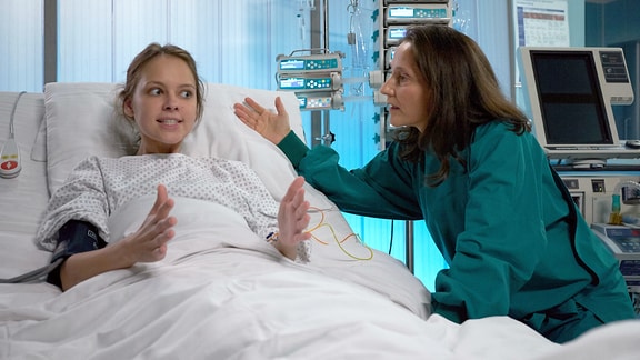 Ein Mädchen in einem Krankenhausbett. Neben ihr sitzt eine Frau. Die beiden scheinen zu diskutieren.