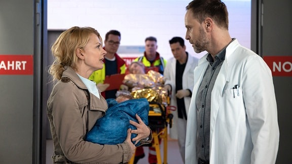 Dr. Ina Schulte (Isabell Gerschke, vorn, li.) hat ein Baby im Arm und steht vor Dr. Kai Hoffmann (Julian Weigend, vorn, re.)