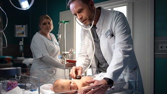 Dr. Kai Hoffmann (Julian Weigend) untersucht ein Baby mit dem Stethoskop.