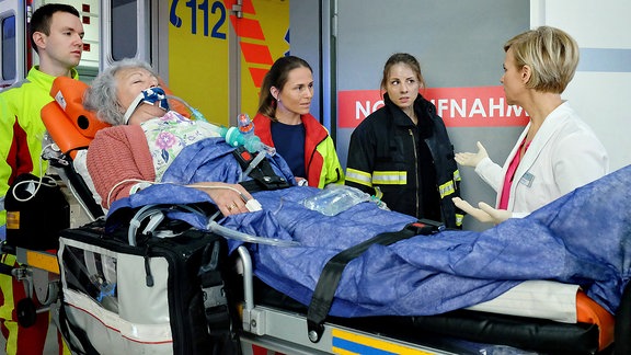 Ein Notarztwagen bringt eine Patientin (Komparsin). Feuerwehrfrau Nina Manzel (Julia Schäfle, 2.v.re.), die Notärztin (Caren Seifert, mi.) und Dr. Kathrin Globisch (Andrea Kathrin Loewig, re.) beraten sich.