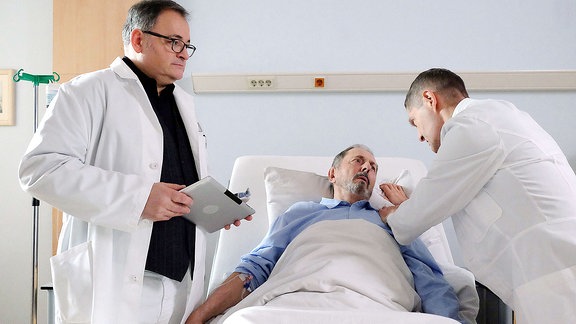 Ein Mann liegt im Bett. Zwei Ärzte kümmern sich um ihn.