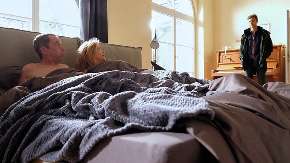 Szene aus "In aller Freundschaft": Eine Frau und ein Mann im Bett, ein junger Mann steht davor.