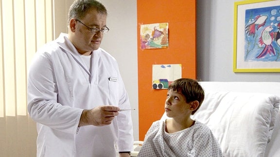 Pfleger Hans-Peter Brenner (Michael Trischan, li.) kontrolliert Noahs (Bruno Renne, re.) Temperatur. Doch Noah versucht zu schummeln. Brenner erklärt dem Jungen, dass er "so" nicht gesund wird.