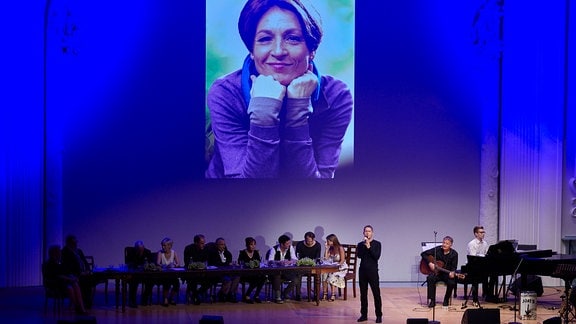 Ein Bild von Hendrikje Fitz wird eingeblendet, davor das Ensemble von In aller Freundschaft auf der Bühne