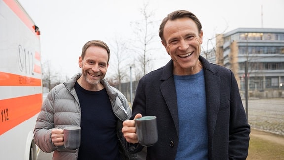 Julian Weigend und Bernhard Bettermann stehen mit Kaffetassen vor der Sachsenklinik