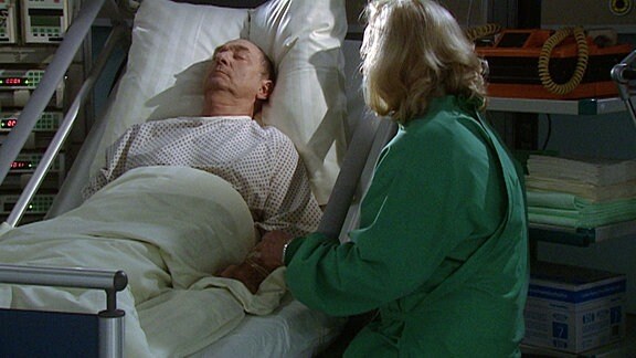 Eine Frau sitzt neben einem Krankenbett in dem ein Mann liegt.