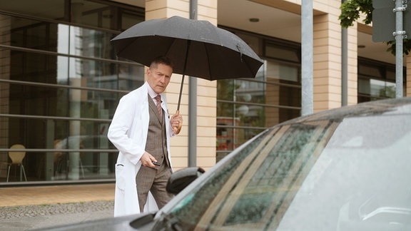Dr. Kaminski steht in seinem Arztkittel und einem aufgespannten schwarzen Regenschirm vor dem Auto von Dr. Demir, einem silbernen Porsche.
