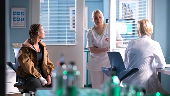 Eine Pflegerin (Miriam) kommt ins Untersuchungszimmer. Die Ärztin (Frau Dr. Globisch) und ihre Patientin (Anna Zübel) sehen sie an.