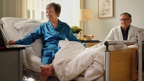 Ein Arzt sitzt traurig neben dem Bett einer fröhlich wirkenden Patientin.