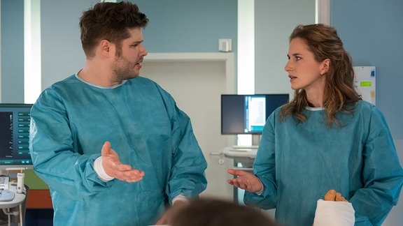 Die Geschwister Fabian und Grit stehen in blauen Krankenhaus-Kitteln in einem Krankenzimmer.