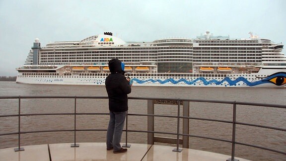 Eine Person steht am Geländer einer Terrasse direkt am Wasser. In einiger Entfernung fährt ein Kreuzfahrtschiff vorbei