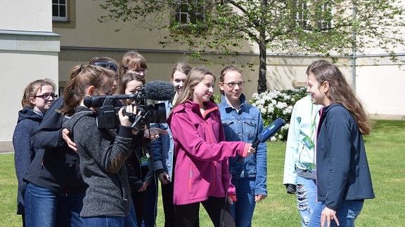 Zwei Mädchen mit Mikrofon und Kamera stehen vor weiteren Mädchen.
