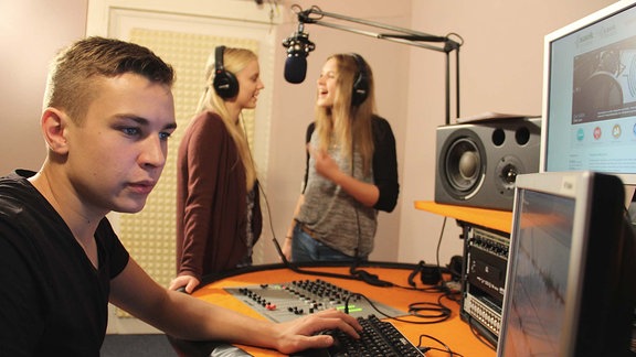 Ein Junge sitzt vor einem Computerbildschirm. Im Hintergrund sprechen zwei Mädchen in ein Mikrofon.