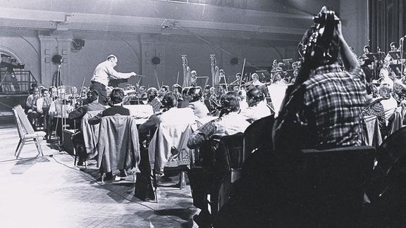 Schwarz-Weiß-Foto des Dirigenten Wolf-Dieter Hauschild und Orchestermusiker von der Seite.