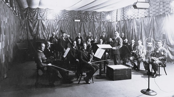 Schwarz-Weiß-Foto von Menschen mit Musikinstrumenten, die ein Orchester bilden und in einem mit Stoff ausgekleideten Raum vor einem Mikofon sitzen.